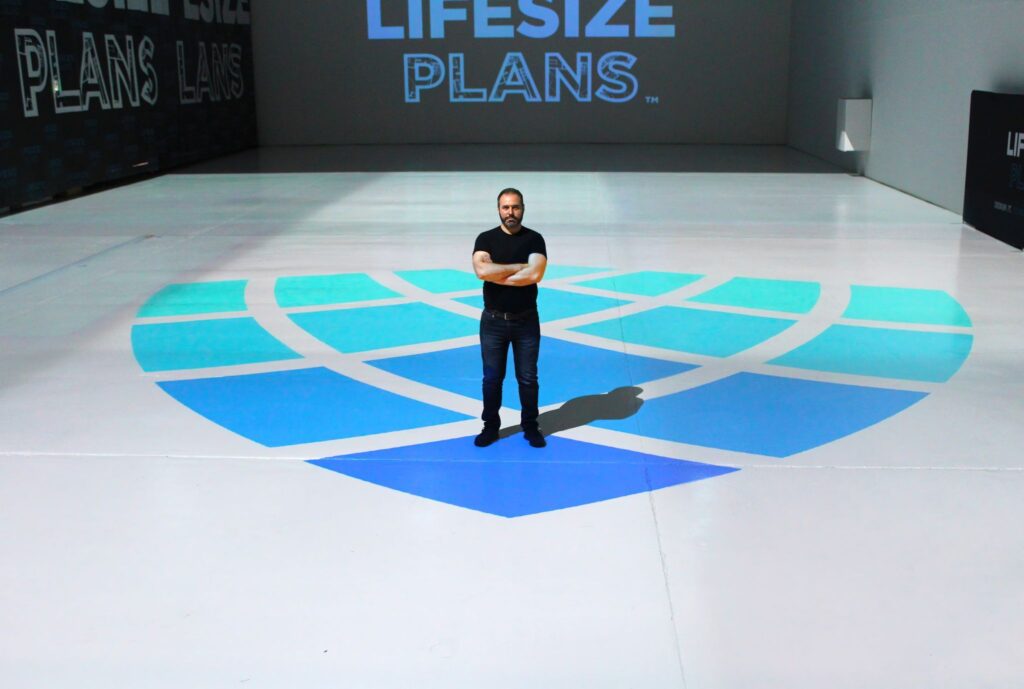Man standing with arms crossed on a stage with a large blue logo and the words "lifesize plans" in the background.