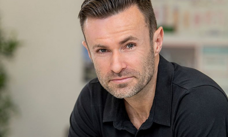 Man with short dark hair and light stubble, wearing a black shirt, sitting indoors while looking intently at the camera